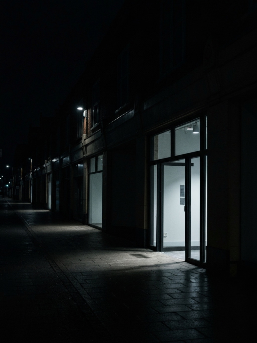 A row of dark empty shopfronts at night with a single unit lit from within