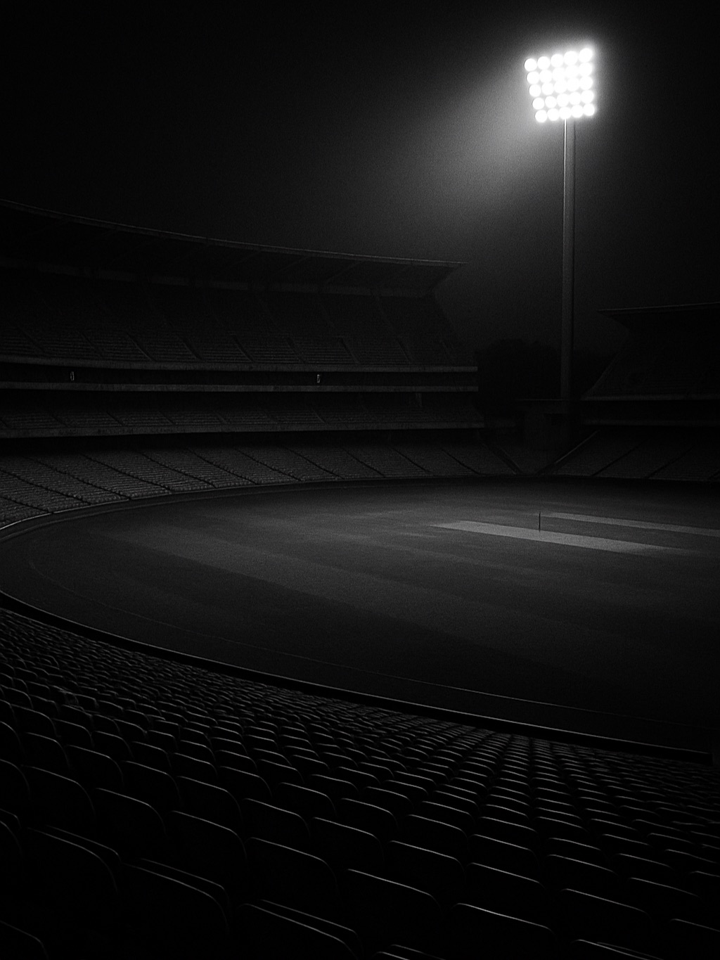 A vast empty cricket stadium at night, one bank of floodlights blazing over empty seats